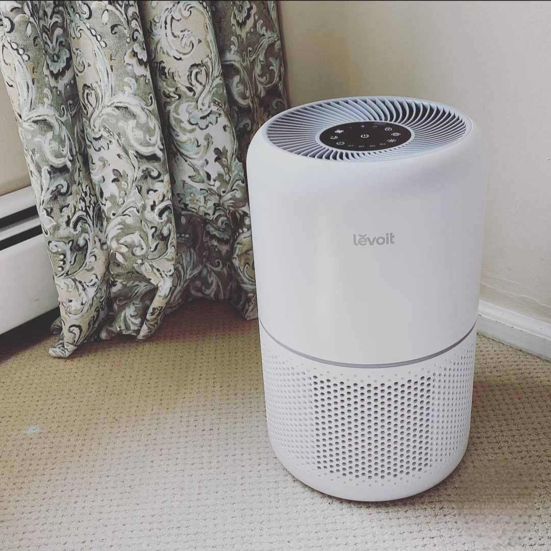 A white air purifier set on top of a carpeted living floor in front of paisley draped curtains.