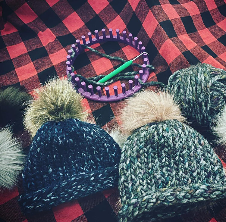 A collection of crochet hats with pom poms  set on top of a red and black plaid blanket with the round loom used to knit the hats.